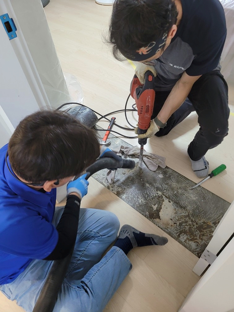 문 앞 바닥을 전동 공구로 뚫는 기사와 옆에서 석션 호스로 먼지를 흡입하는 기사의 모습